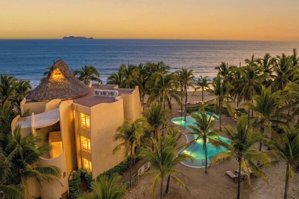 A resort with a pool, palm trees, and a view of the ocean at sunset.