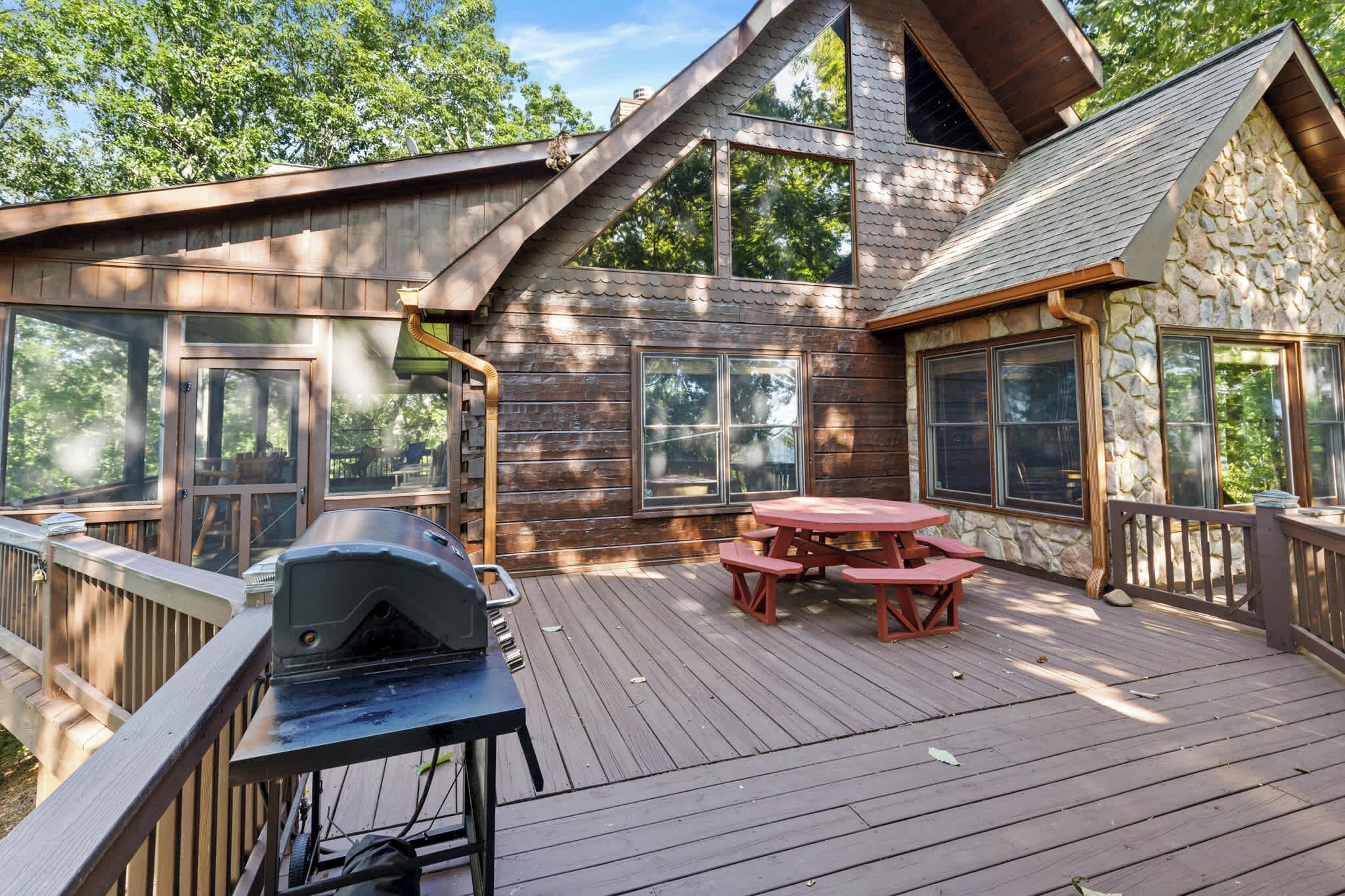 A wooden deck with a barbecue grill and picnic table, surrounded by stone walls and large windows.