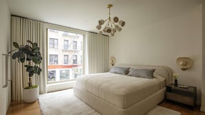 Ein Schlafzimmer mit einem großen Bett, einer Zimmerpflanze, einem Nachttisch und Blick auf Gebäude.