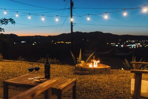 Et udendørs siddeområde med et bord, to glas rødvin og en flaske, foran en pejse og lyskæder.