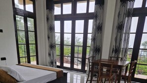 Un dormitorio con cama, una mesa de comedor con sillas, ventanas amplias y vistas al verdor exterior.
