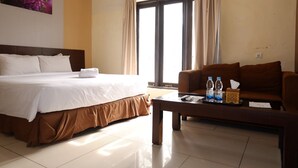 A hotel room with a large bed, a brown sofa, a coffee table with water bottles, and a window with curtains. - De Nusantara Hotel Syariah (Medan)