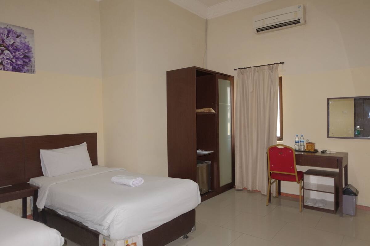A hotel room with two beds, a desk, a chair, a wardrobe, and an air conditioning unit.