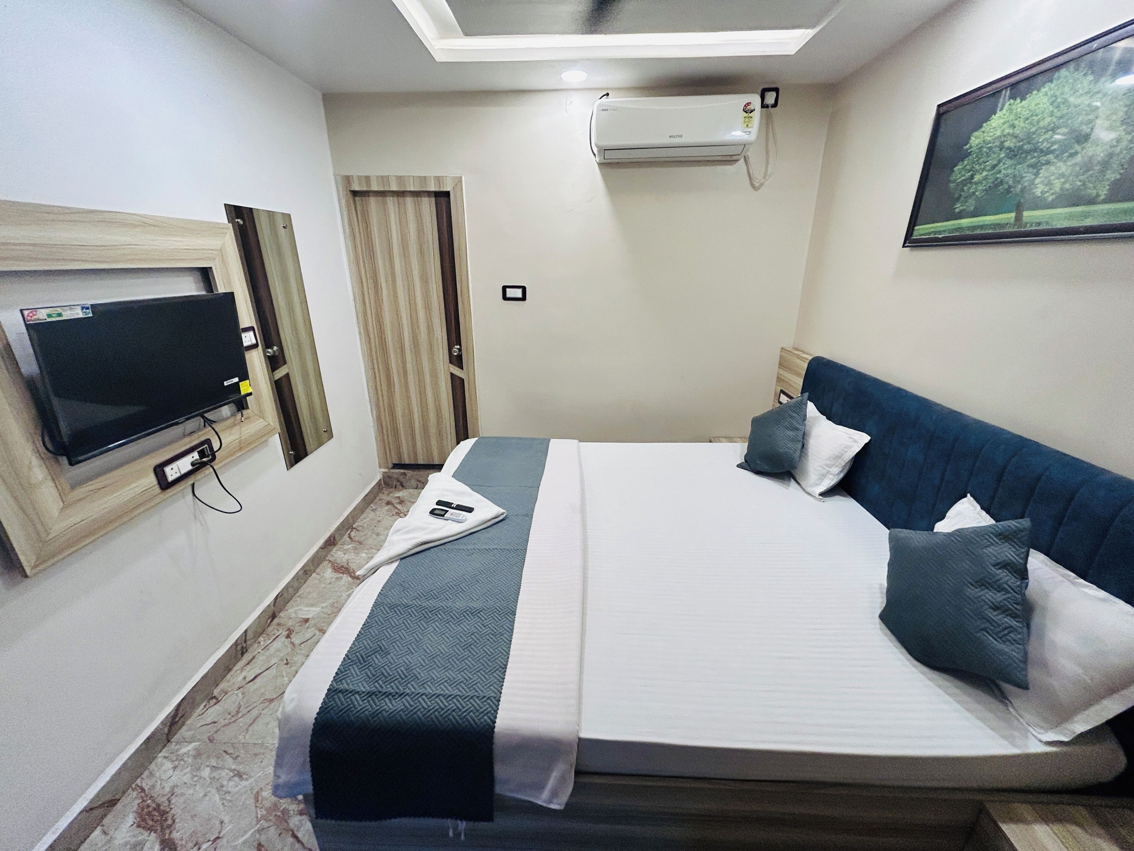 Una habitación de hotel con cama, televisor de pantalla plana, equipo de aire acondicionado, un cuadro enmarcado en la pared y una puerta.
