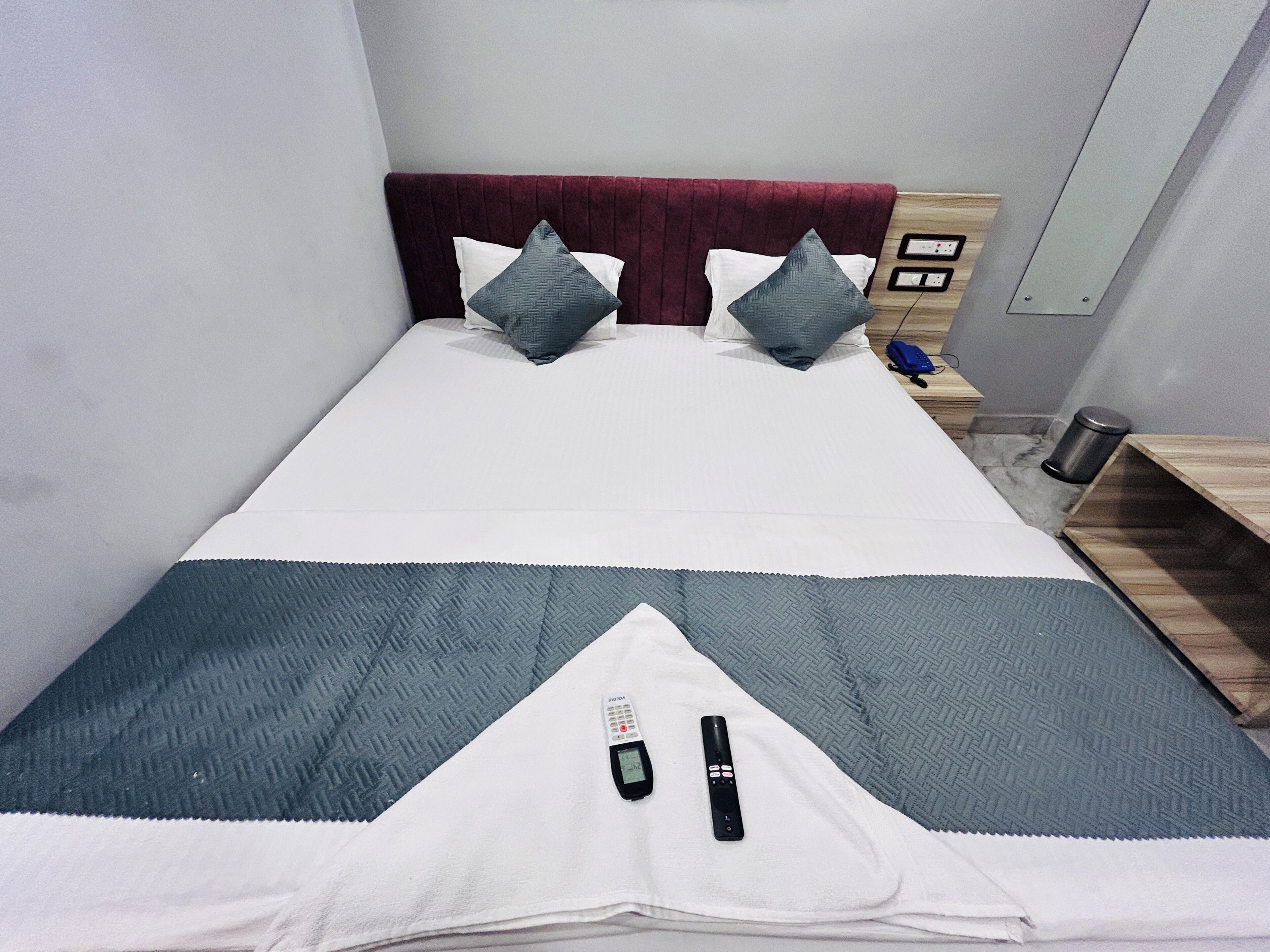Una cama con una manta blanca y gris, dos almohadas, un control remoto y una mesita de noche con una lámpara.