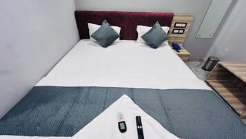 Una cama con una manta blanca y gris, dos almohadas, un control remoto y una mesita de noche con una lámpara.