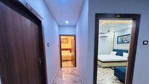 Ein Hotelgang mit Marmorboden, einer Holztür links und einem Zimmer mit Bett und Gemälde, das durch eine geöffnete Tür sichtbar ist.