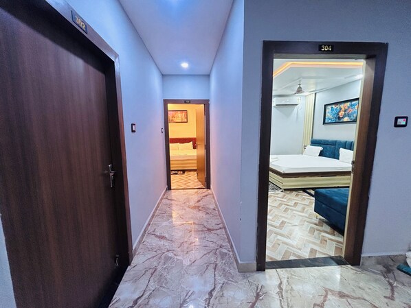 A hotel corridor with a marble floor, a wooden door on the left, and a room with a bed and a painting visible through an open door. - Hotel Dwarka Palace (Darbhanga)
