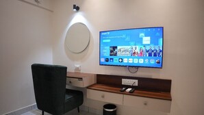 A modern hotel room with a flat-screen TV mounted on the wall, a wooden console table, a black trash bin, and a dark green chair. - AK CASTLE SUITES (Bengaluru)