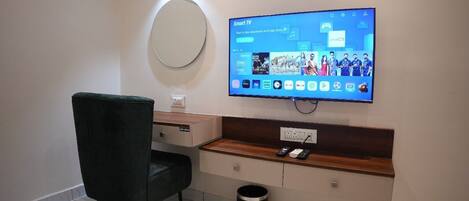 A modern hotel room with a flat-screen TV mounted on the wall, a wooden console table, a black trash bin, and a dark green chair.