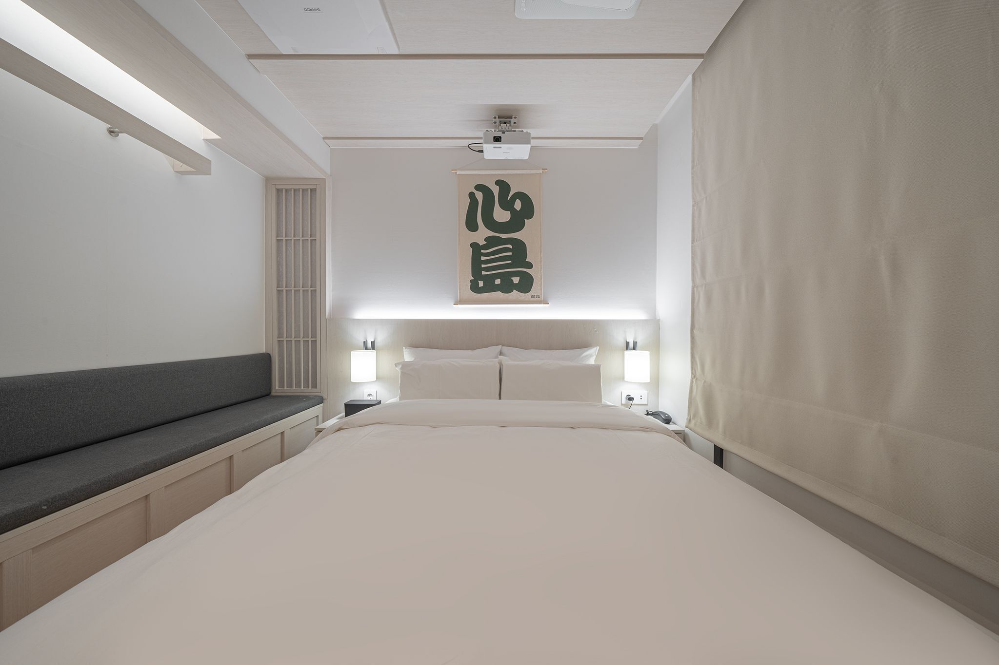 A minimalist bedroom with a large bed, bedside lamps, a wall-mounted projector, and a hanging sign with Chinese characters.