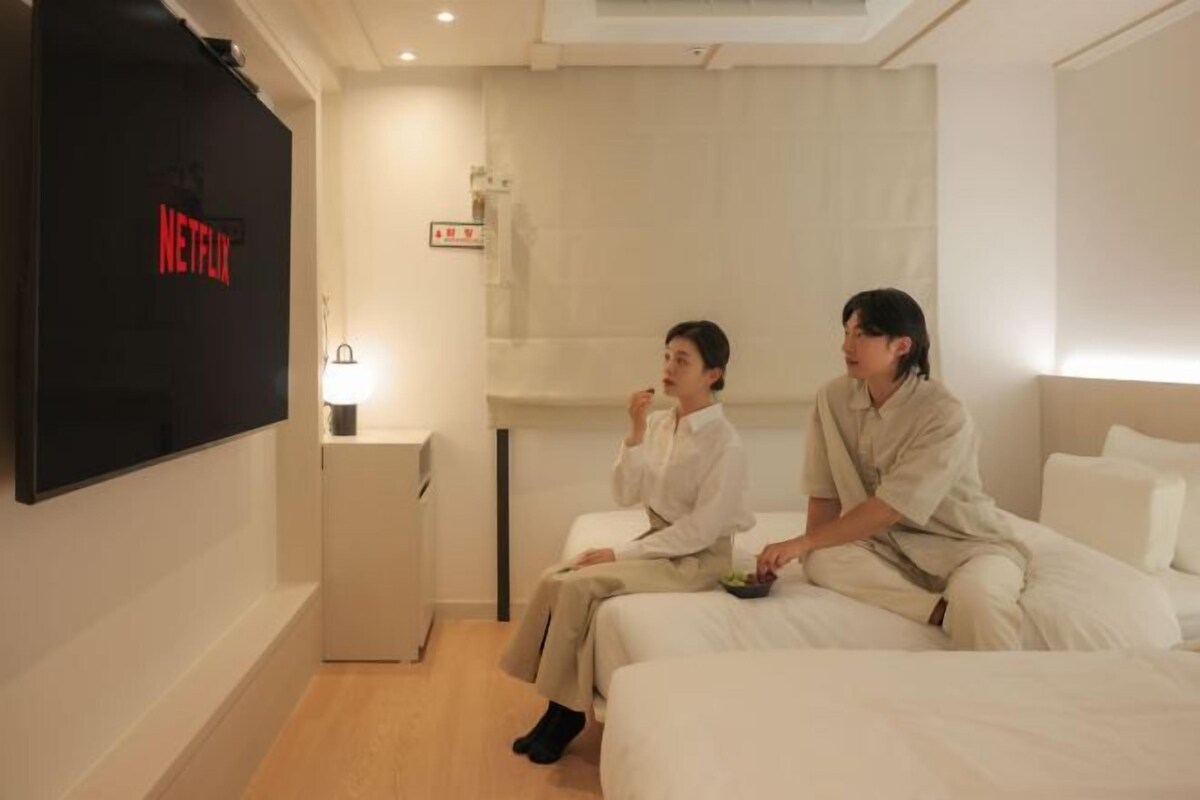 Una habitación de hotel con una cama, una mesita de noche, un televisor mostrando el logotipo de Netflix y un mueble pequeño.