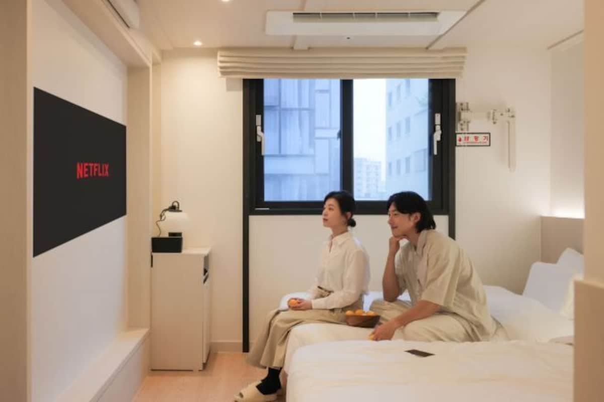 Habitación de hotel con cama, mesita de noche pequeña, ventana con vista a edificios y televisor de pared mostrando el logotipo de Netflix.
