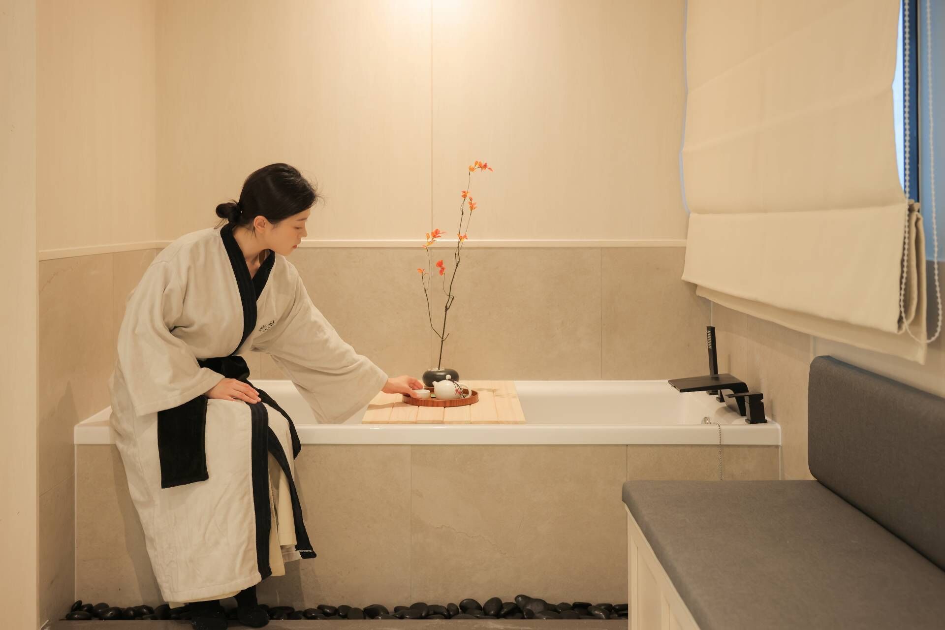 Eine Person in einem weißen Bademantel bereitet ein Teeset in einem modernen Badezimmer mit einer Badewanne, einer grauen Sitzbank und einem kleinen Tisch mit einem Blumenstrauß vor.