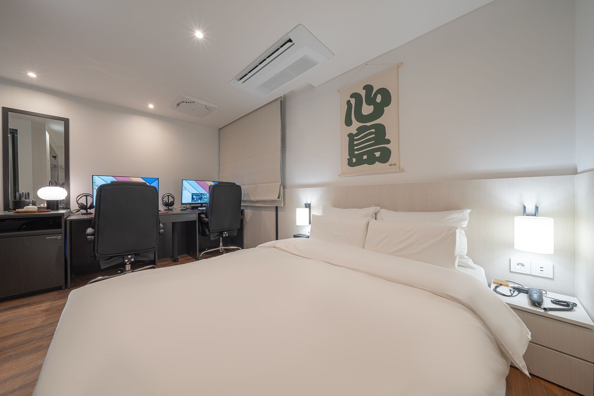 A hotel room with a large bed, a desk with two monitors, a chair, and a desk lamp.