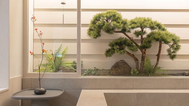 Un jardín interior minimalista con un bonsái, una mesita con un florero y vista a un edificio.