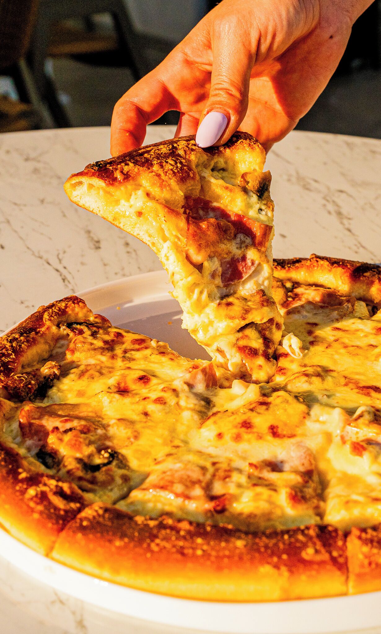 Uma mão segurando uma fatia de pizza com queijo e acompanhamentos, sobre um prato branco.