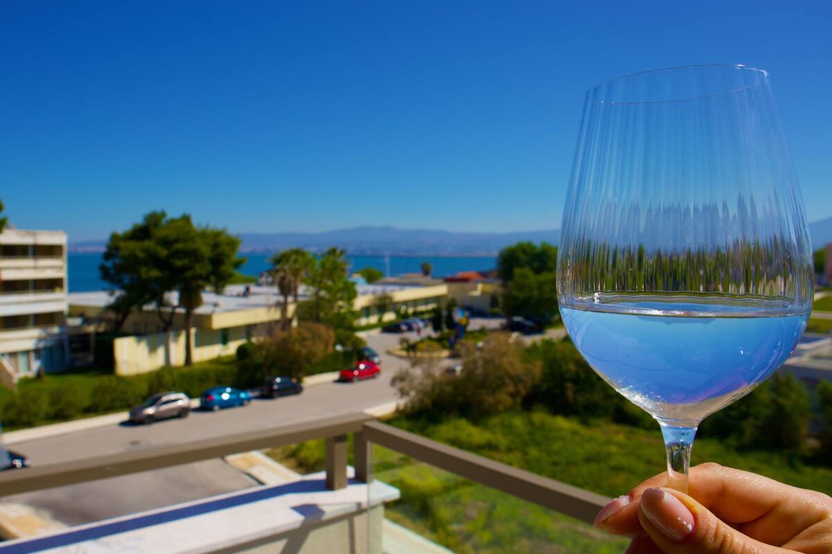 Una mano sostiene un vaso de vidrio transparente con un líquido azul, con vistas a una zona residencial que incluye casas, una carretera y una masa de agua bajo un cielo azul despejado.
