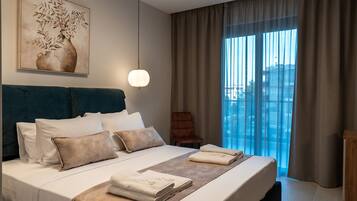 A hotel room with a large bed, a view of the city through the window, a painting on the wall, and a hanging light fixture.