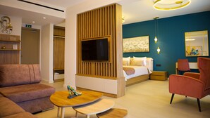 Habitación de hotel moderna con sofá, una pequeña mesa de madera, televisor montado en un panel de madera, cama con una pared de acento azul y una silla.