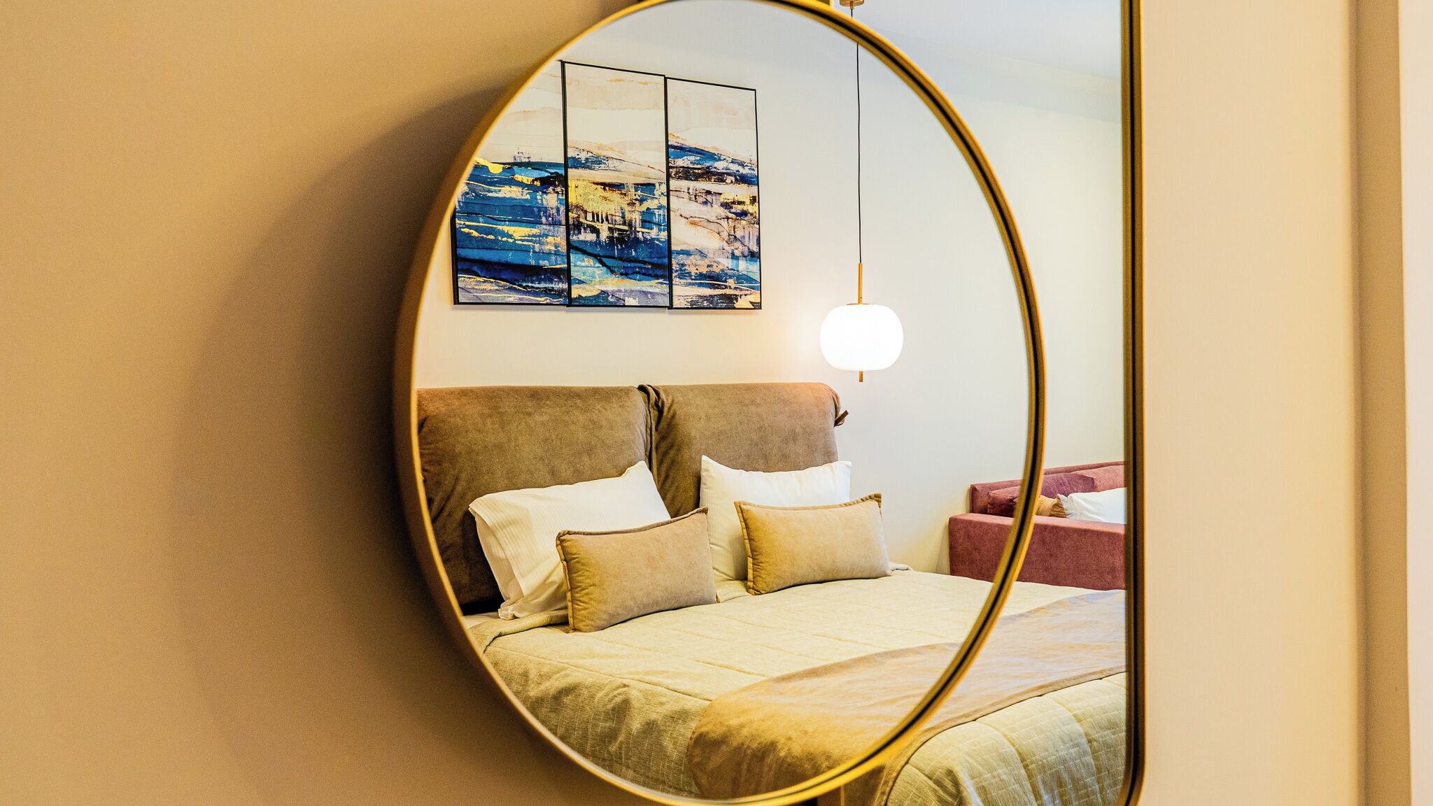 Un dormitorio con un espejo ovalado grande, una cama con ropa de cama color beige y una lámpara de pared.