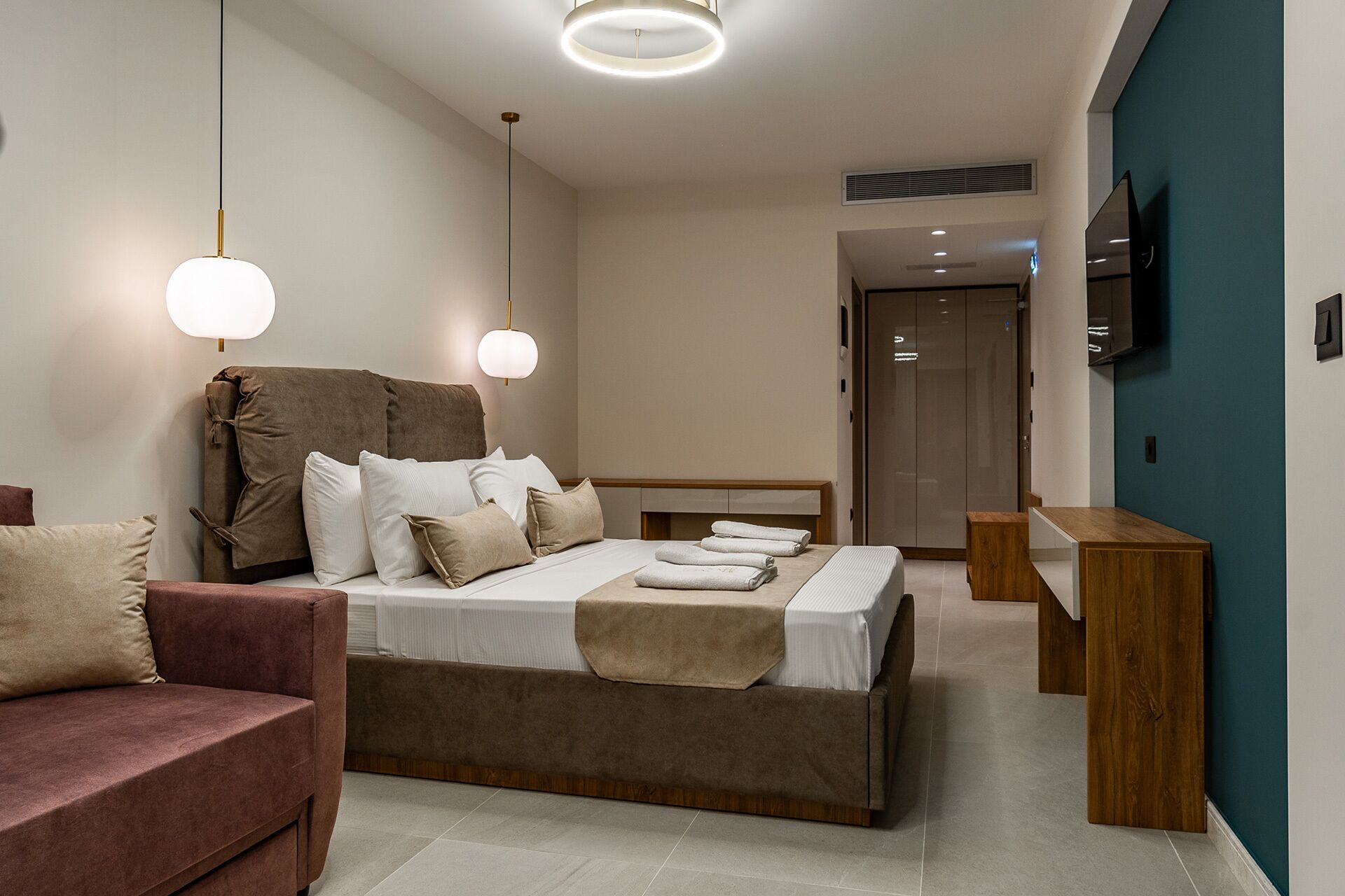 A modern hotel room with a large bed, a sofa, a wooden side table, and a TV mounted on the wall.