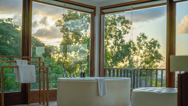 Une salle de bain avec une baignoire, un banc en bois et une grande fenêtre donnant sur des arbres et un ciel nuageux.