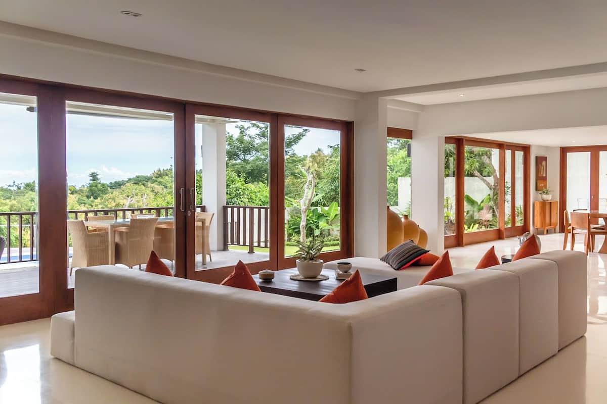 A spacious living room with a large white sofa, a coffee table with a plant, and a view of lush greenery outside.