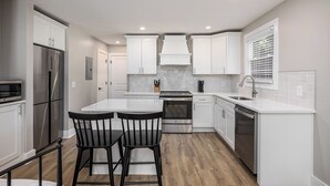 Una cocina moderna con armarios blancos, electrodomésticos de acero inoxidable, una isla central con dos sillas negras y una ventana con persianas.