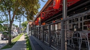 Un espacio para comer al aire libre con sillas y mesas de metal, faroles rojos y una acera con coches estacionados.