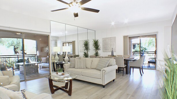 A spacious living room with a large sofa, a coffee table, and a dining area with chairs and a table. There is a ceiling fan, a large mirror, and a view of the outdoors through a sliding glass door. - 3BR condo near WestWorld (Scottsdale)