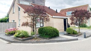 Un edificio residencial de dos pisos con fachada de ladrillo, techo a dos aguas y un jardín delantero con diversas plantas y un árbol pequeño.