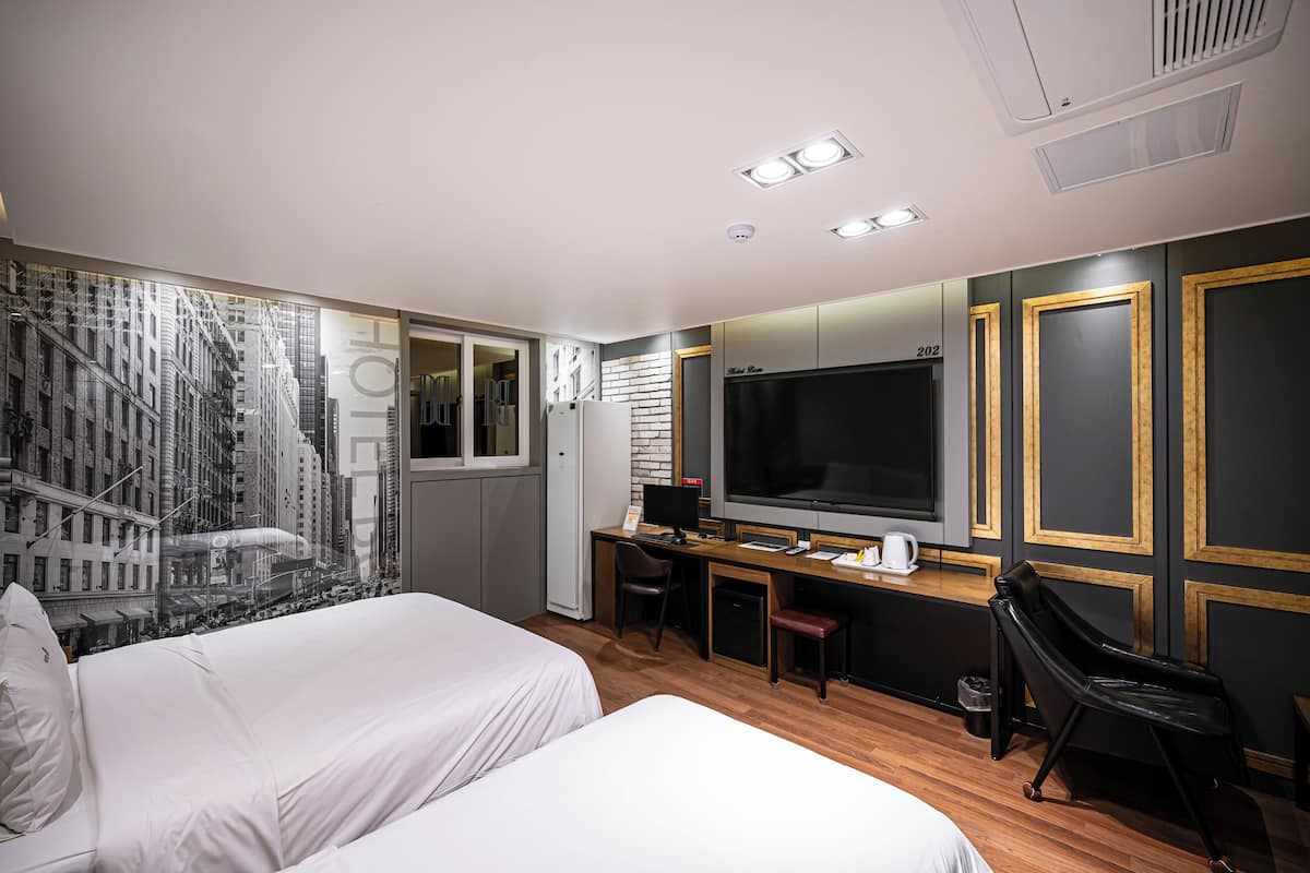 A hotel room with two beds, a desk with a computer, a TV, and a large window with a cityscape view.