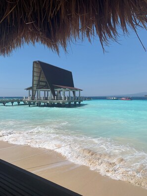 Eine Strandstruktur mit einem großen, vorspringenden Dach, einer Pier, die sich in das türkisblaue Meer erstreckt, und einem Boot im Hintergrund.