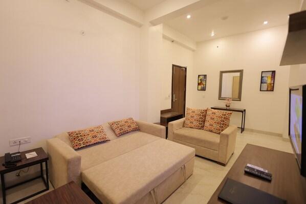 A modern hotel room with two beige sofas, a small wooden table, a TV, and a desk with a phone and charger. - Hide Away Studios (Mathura)