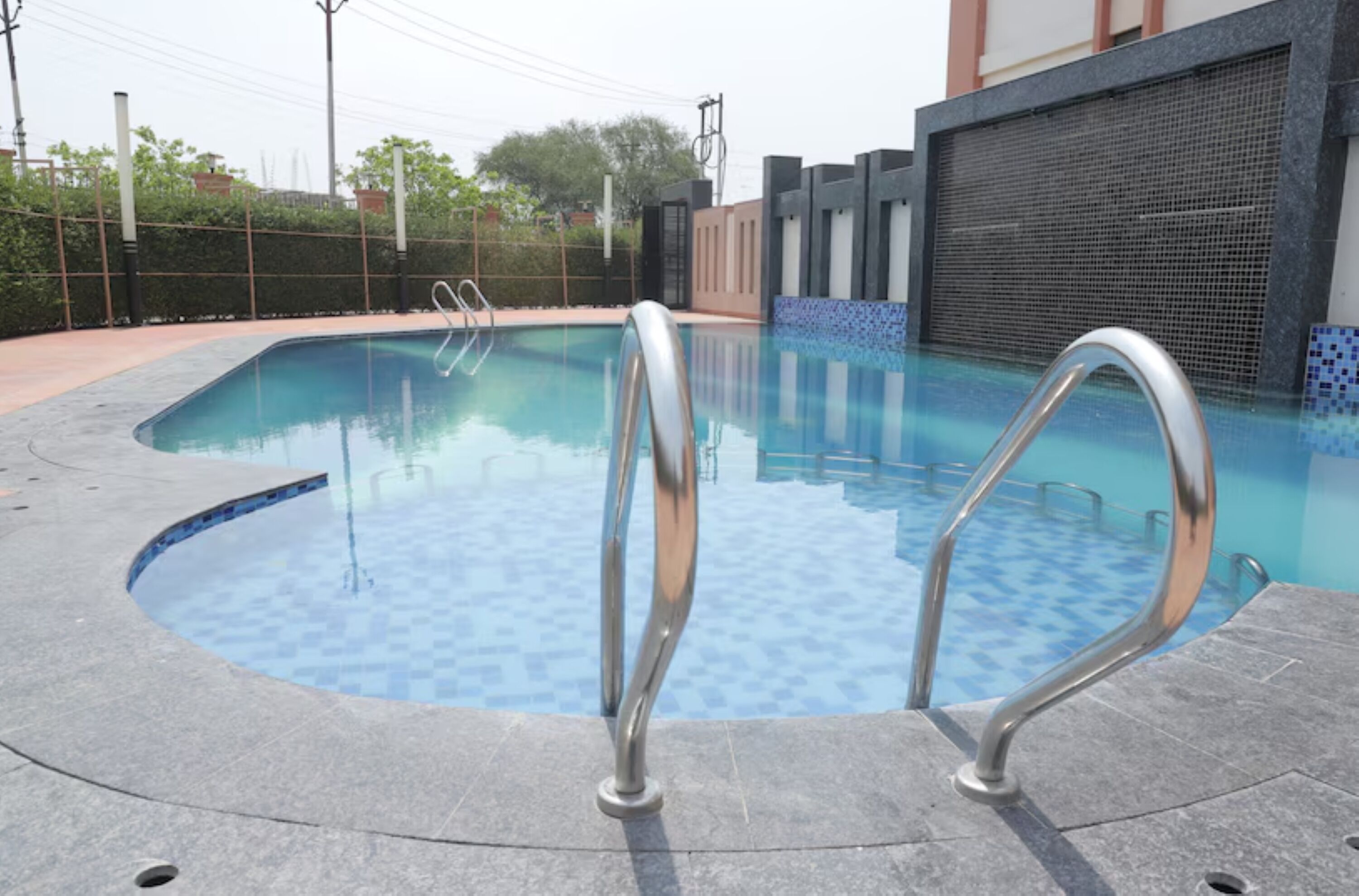 A swimming pool with metal handrails, surrounded by a tiled deck and a fence.