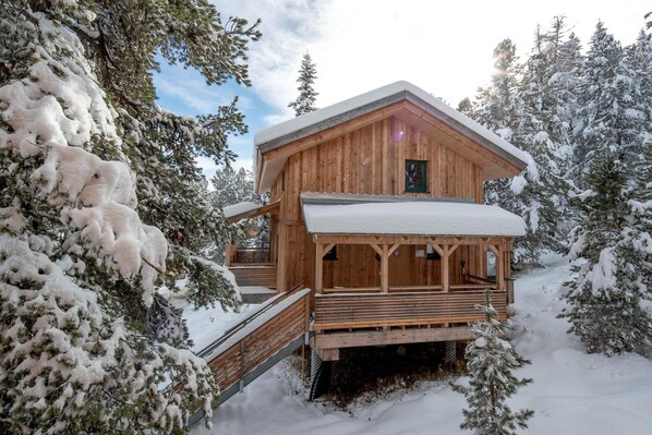 A wooden cabin with a covered porch and a staircase leading up to it, surrounded by snow-covered trees.