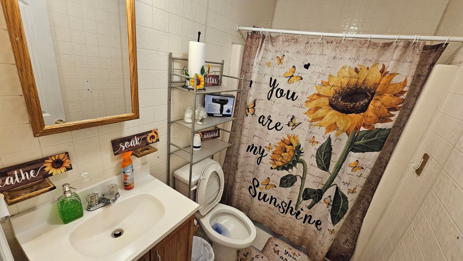 A bathroom with a toilet, sink, and shelves with toiletries. There is a shower curtain with a sunflower and butterfly design.