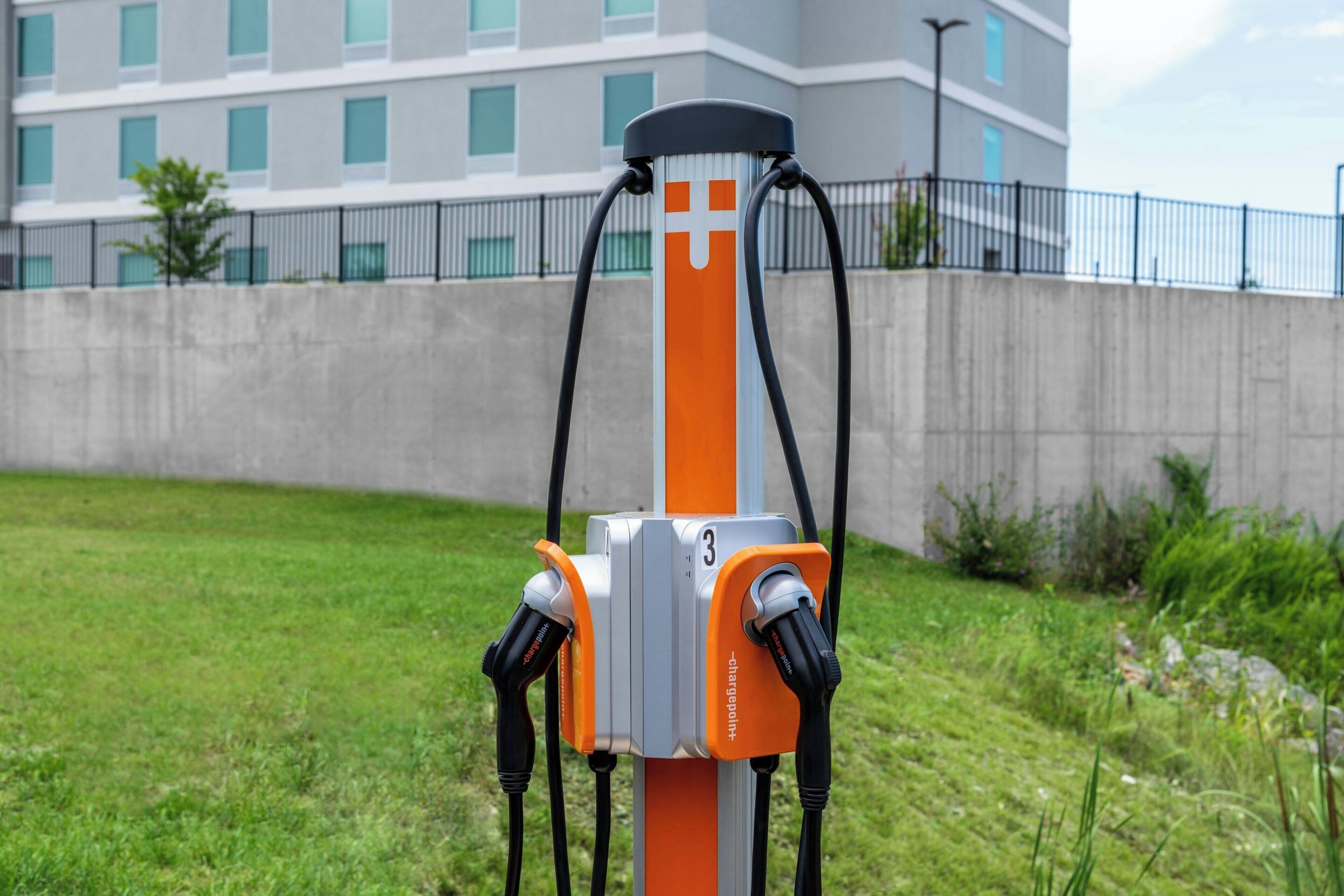 Una estación de carga para vehículos eléctricos con dos conectores, frente a un edificio y una zona verde.