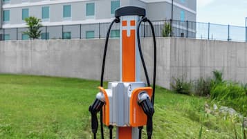 Una estación de carga para vehículos eléctricos con dos conectores, frente a un edificio y una zona verde.