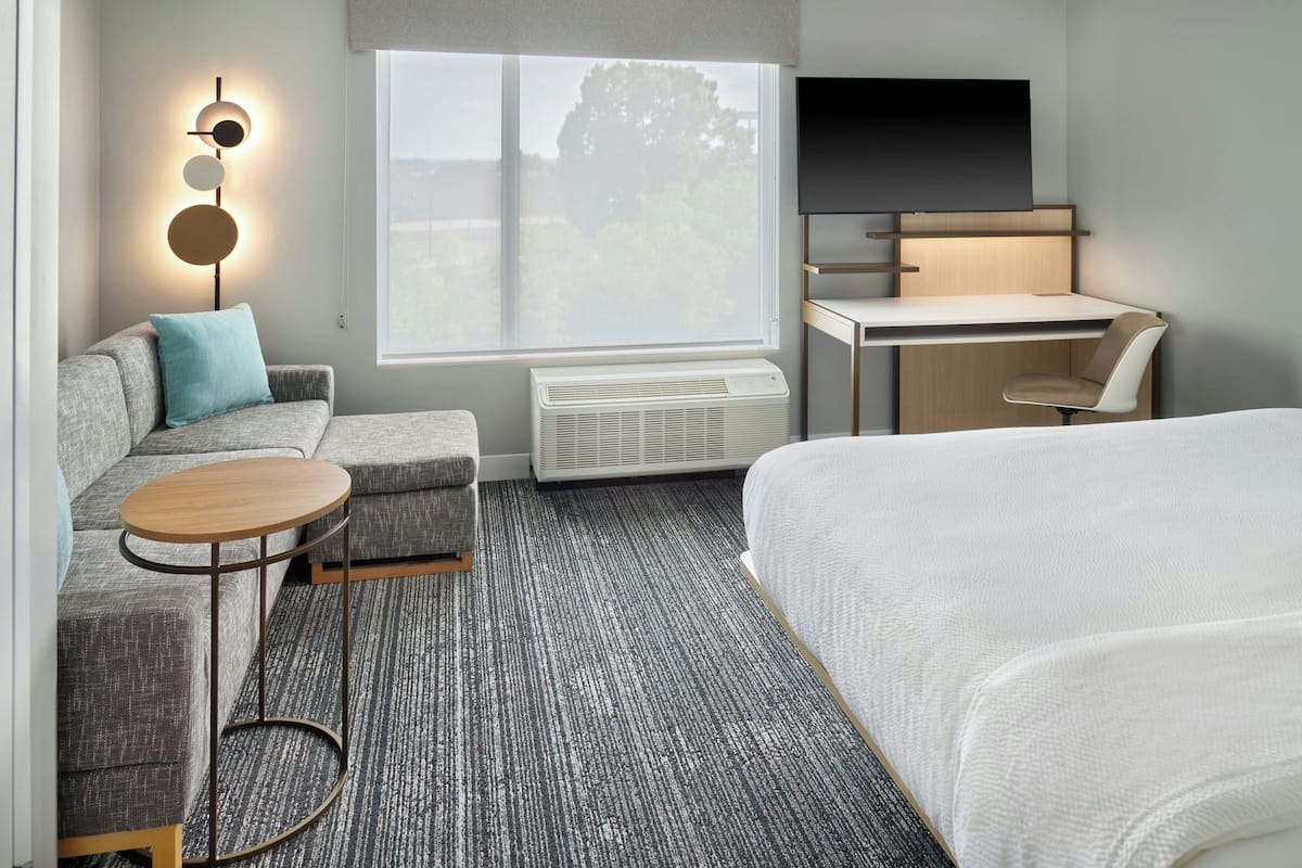 A hotel room with a bed, a desk, a chair, a sofa, a small round table, and a wall-mounted light fixture.