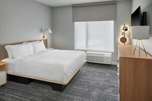Habitación de hotel moderna con una cama grande, mesitas de noche, un escritorio, una silla, un televisor y una ventana con persianas.