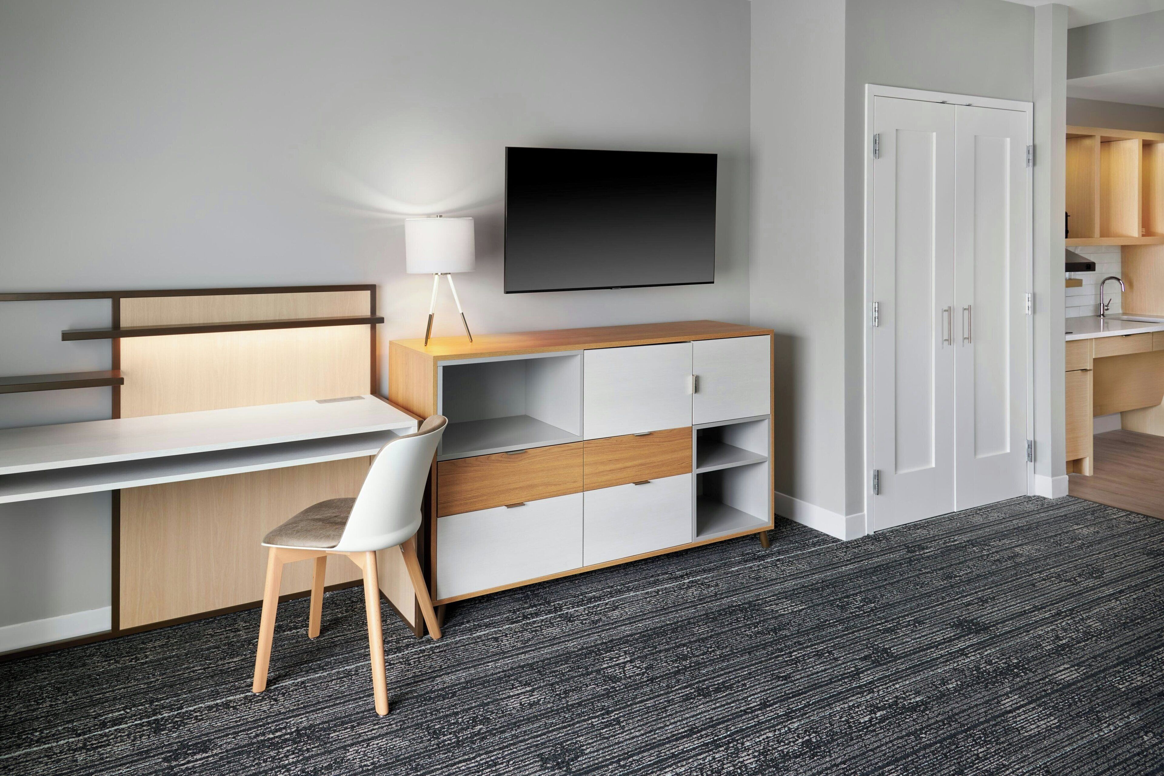 A modern room with a desk, chair, TV, wardrobe, and kitchenette.