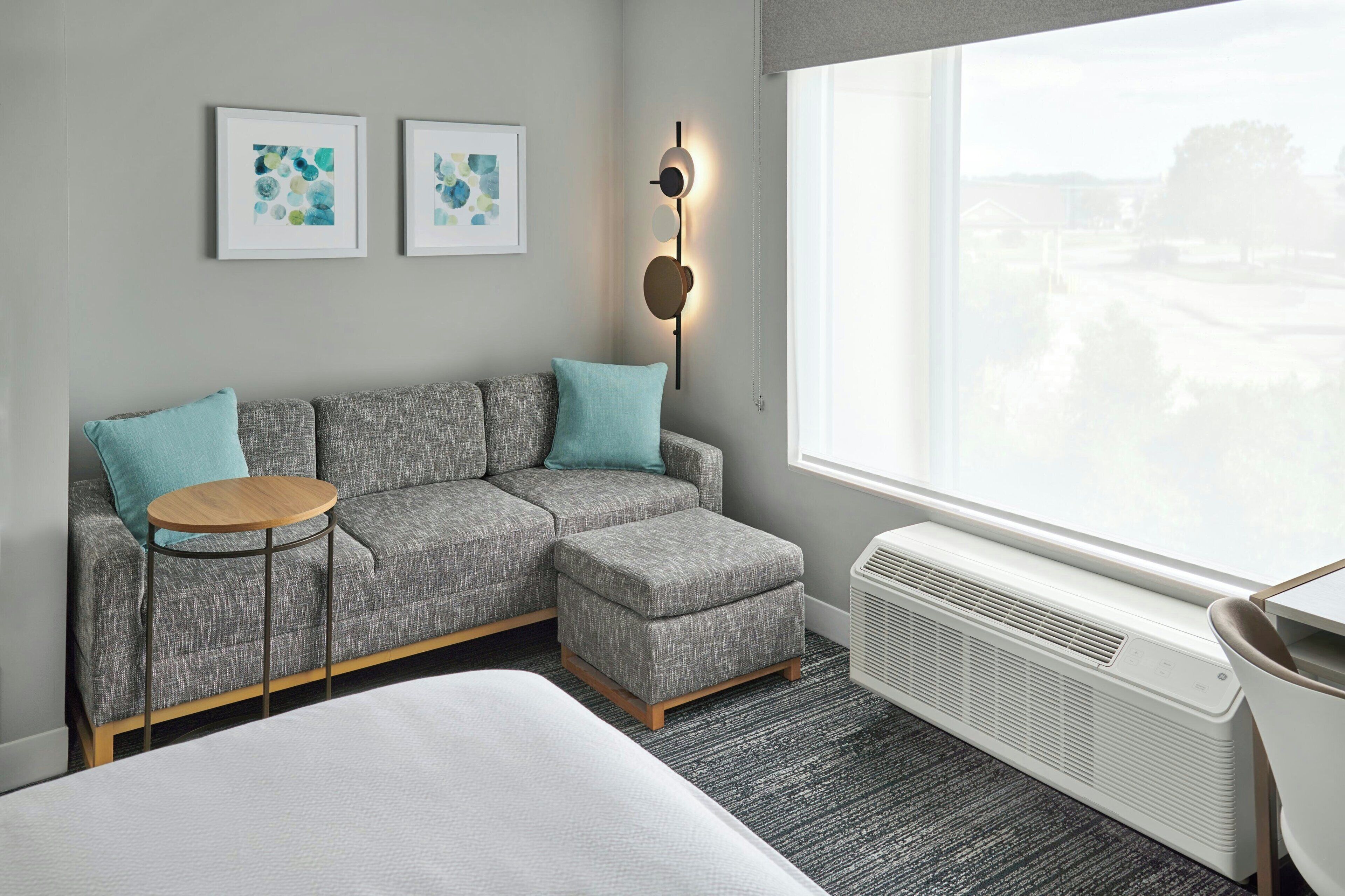 Een moderne hotelkamer met een grijze bank, een klein rond tafeltje, een bed en een groot raam met uitzicht.
