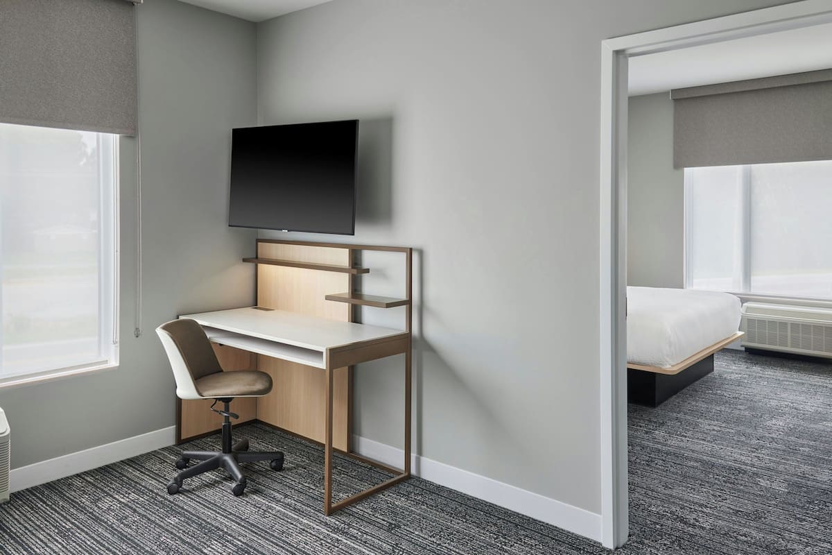 A hotel room with a desk, chair, TV, bed, and large windows.