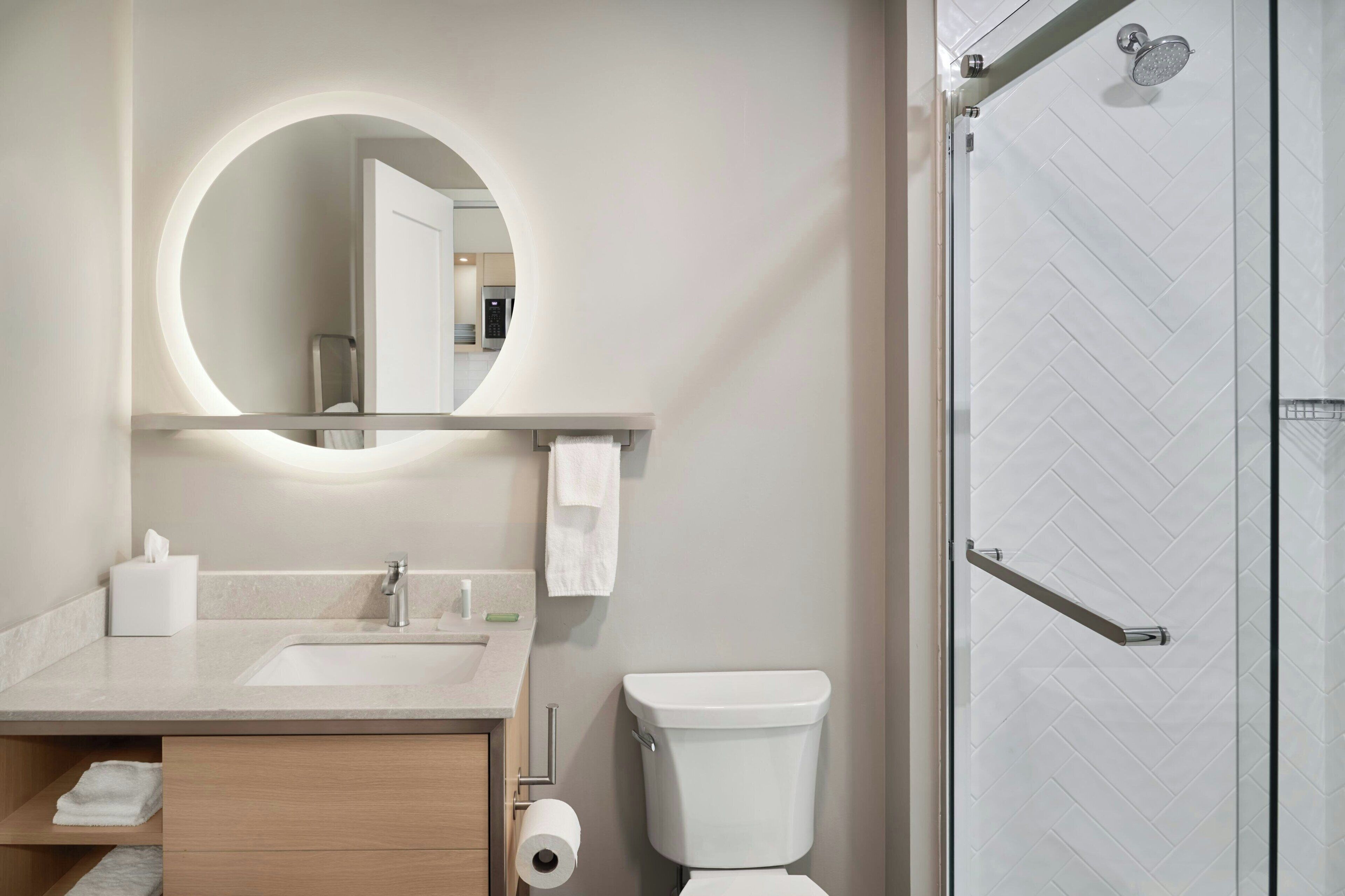 A modern bathroom with a round mirror, two sinks, a toilet, and a glass shower enclosure.