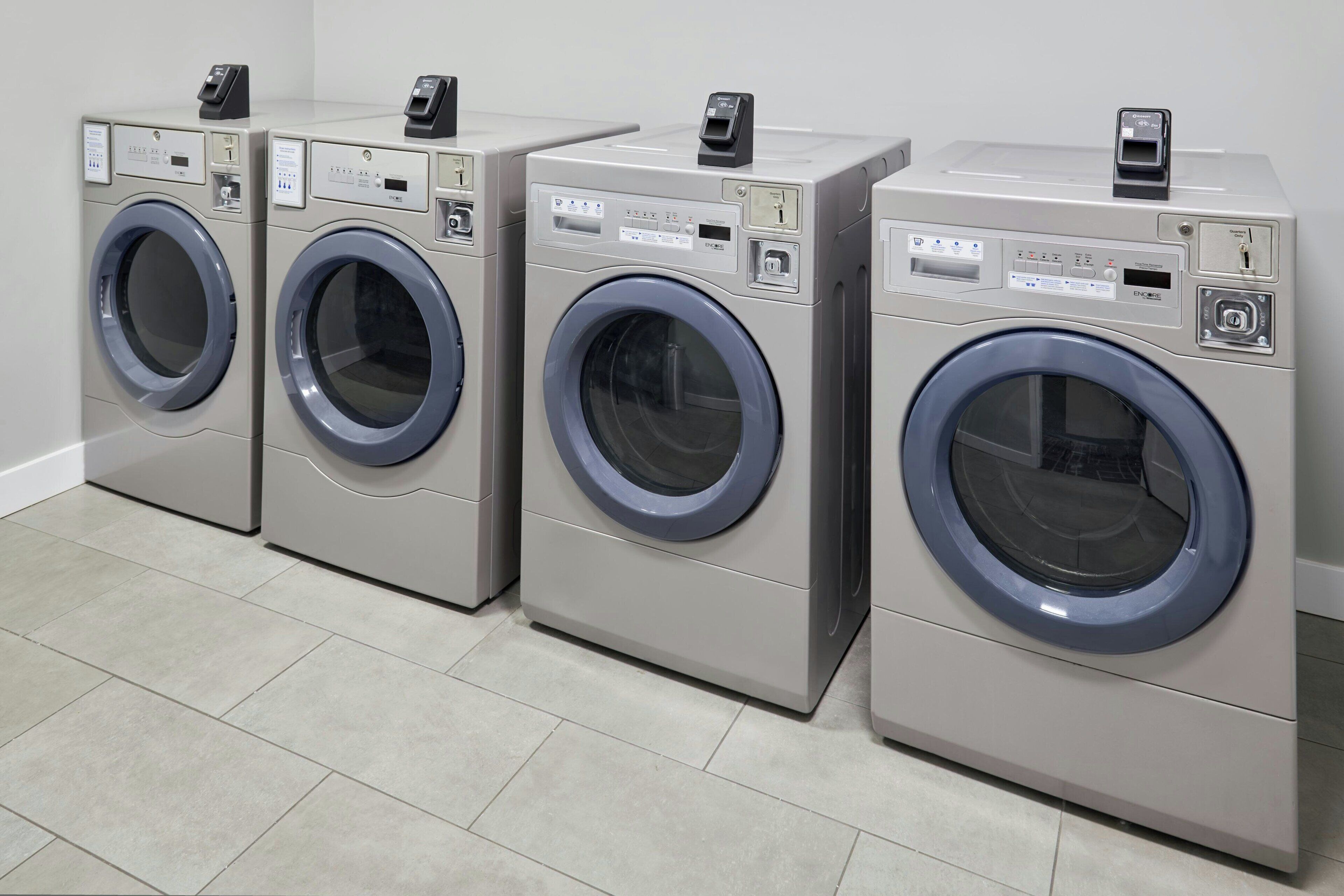 A row of four identical front-loading washing machines in a commercial setting.
