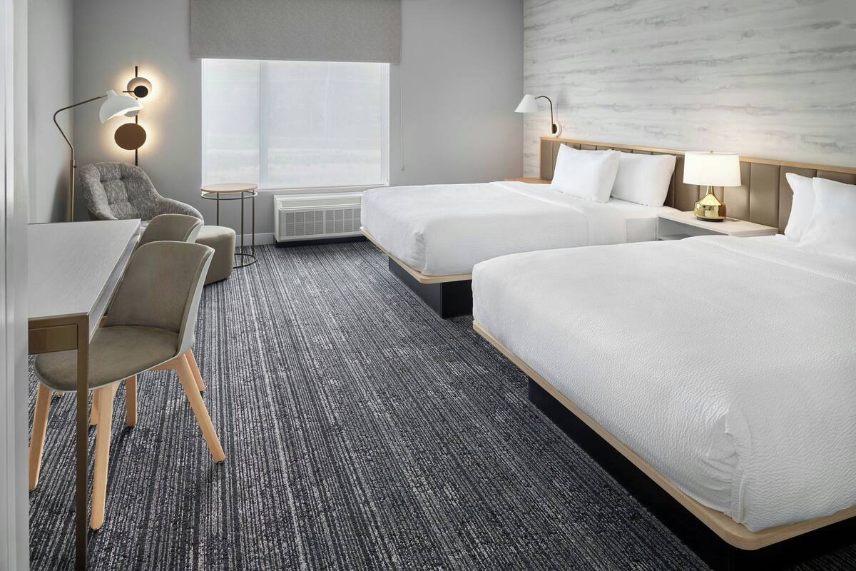 A hotel room with two beds, a desk, a chair, and a lamp.