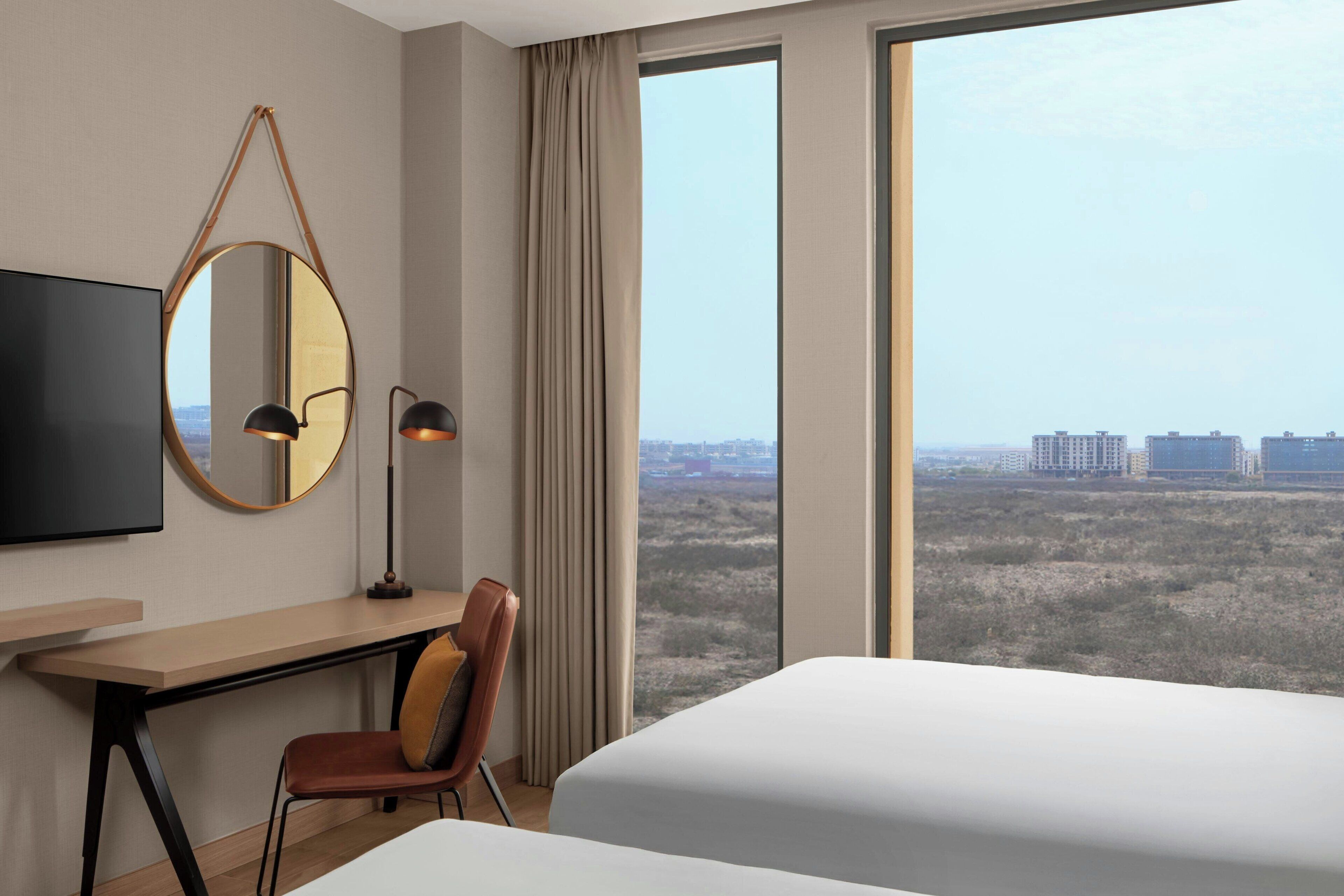 A hotel room with a bed, a desk, a chair, a TV, a mirror, and a large window offering a city view.