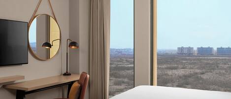 A hotel room with a bed, a desk, a chair, a TV, a mirror, and a large window offering a city view.